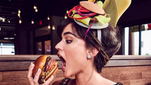 Woman in a navy dress and burger-inspired bonnet eating a burger.