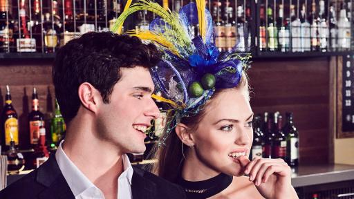 Man in black suit leaning against the bar, holding a margarita. A woman in a black dress wearing a margarita-inspired bonnet leans next to him.