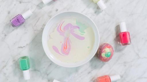 Nail polish and one egg surround a bowl of what with some nail polish on top. The egg shows the results of dipping in the water, a marbling technique.