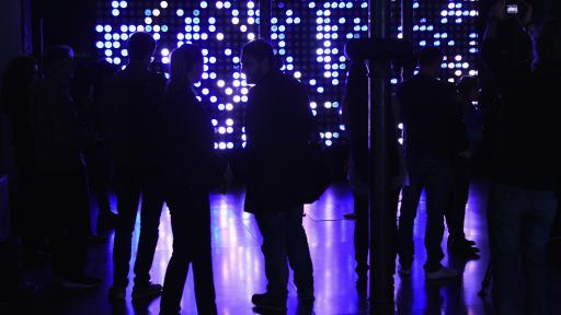 People standing and talking in a dark room lit only by purple lights on the far wall.
