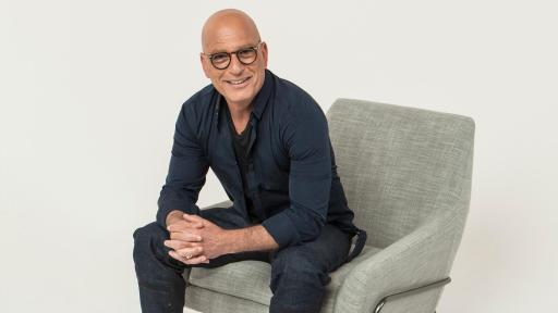 Howie Mandel in glasses sitting on a chair with red shoes among a plain gray background