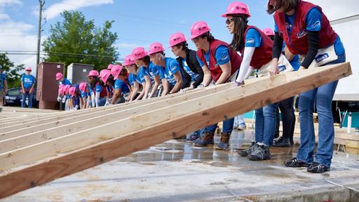Women in hardhats working together to lift a latticework structure of wood for a wall.