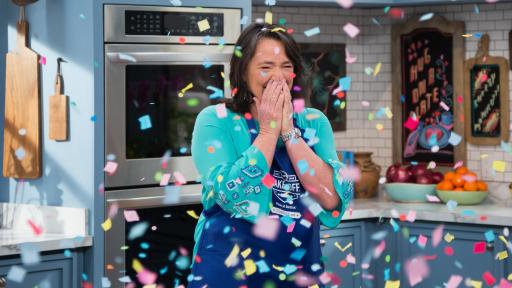 A very happy Amy Nelson stands behind a kitchen counter being showered with confetti.