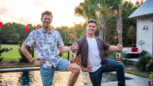 Adam Devine and Tyler Tills strike a pose with a bottle of rum on their knees in front of a pool.