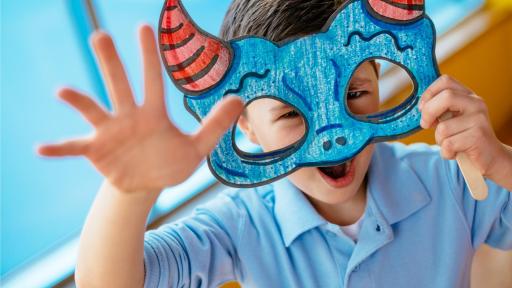 A young boy holds up a monster mask that he has colored with crayons.