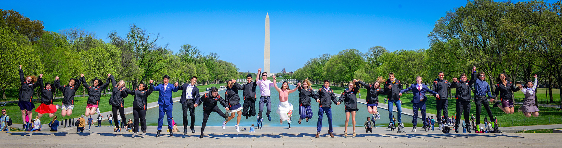 A line of young people, holding hands in mid-jump. They are lined up in front of the 
Lincoln Memorial Reflecting Pool with the Washington monument in the background.