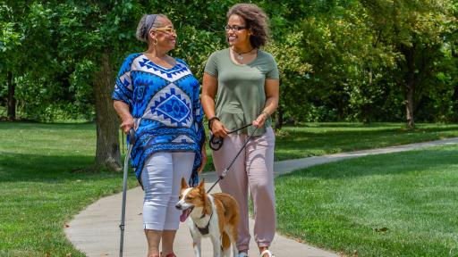An elderly woman and younger woman walking a dog through a grassy park, smiling at each other