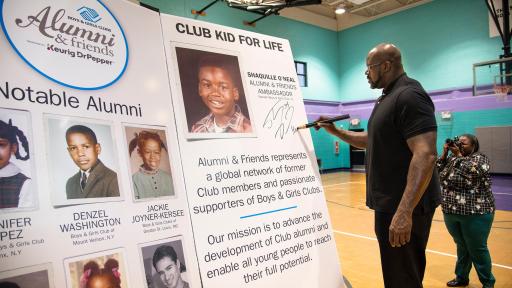 Shaquille O&rsquo;neal autographing the presentation poster in a blue gymnasium.