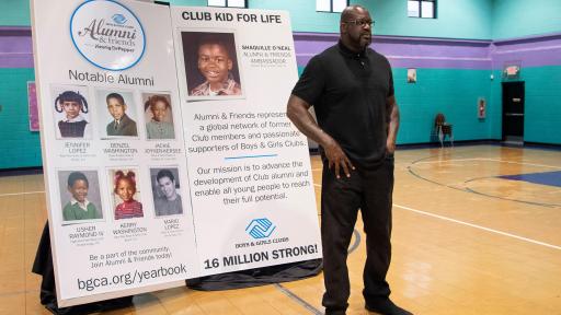 Shaquille O’neal standing in front of a large presentation poster in a blue gymnasium.