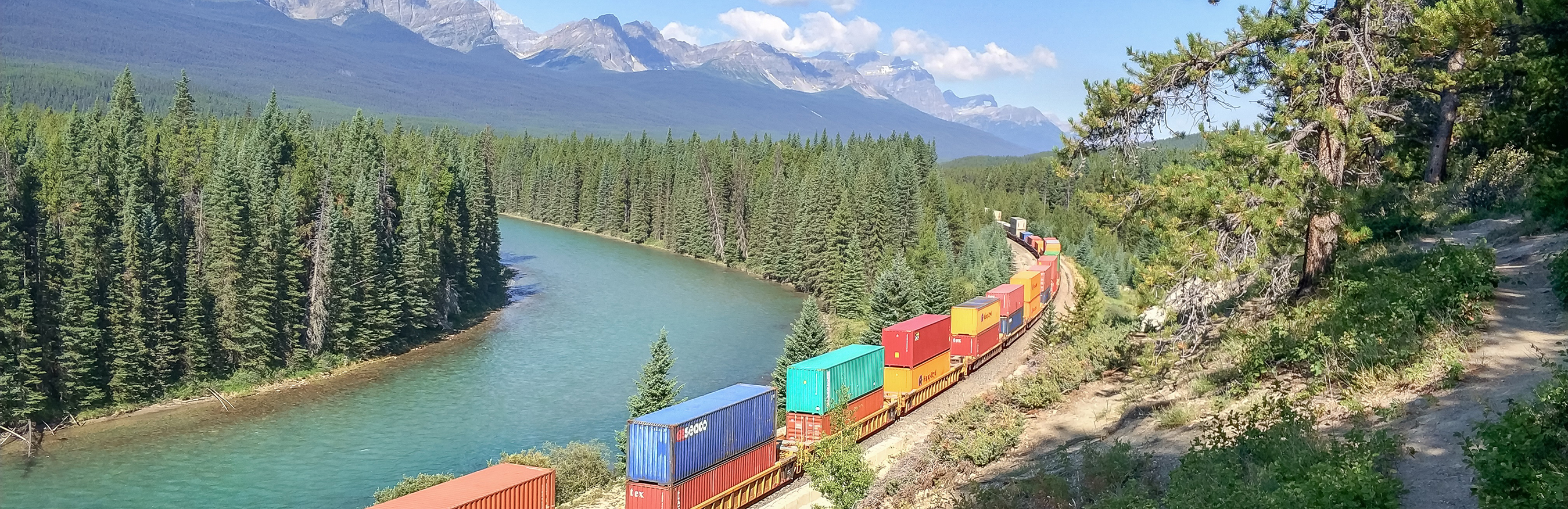 Train taking a turn along a riverbank through a forest of pine trees, with a backdrop of mountains, sky, and clouds.