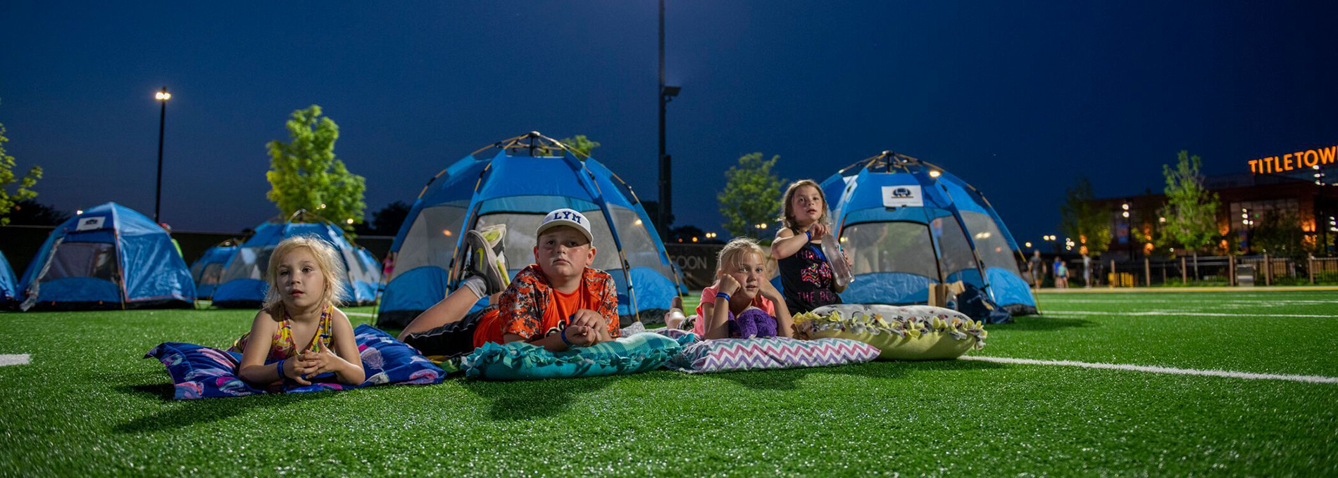 Four children on a sports field with tents in the background.