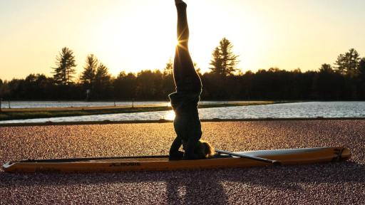 Becca does a headstand on a surfboard in a lake.