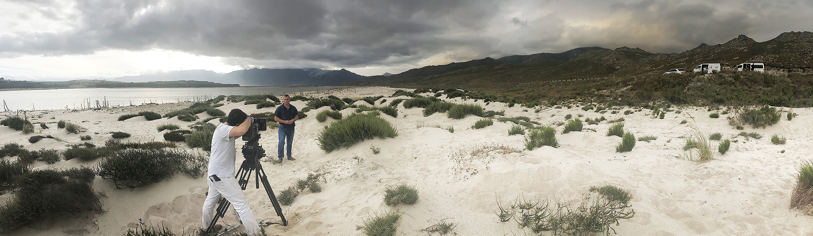 Man filming another man talking on a shore under thick overcast