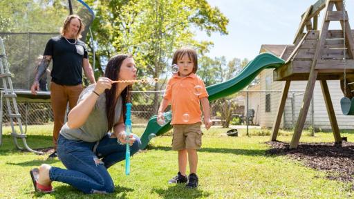 Family playing in a backyard. The mother is blowing bubbles for her small child.