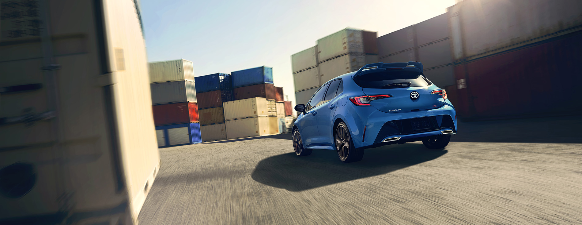 A blue Toyota Corolla drives away, surrounded by shipping containers.