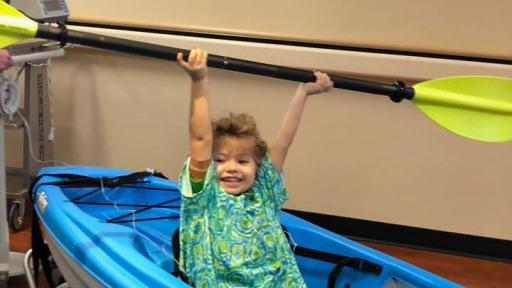 Child wearing a hospital gown, playing in a kayak inside a hospital room