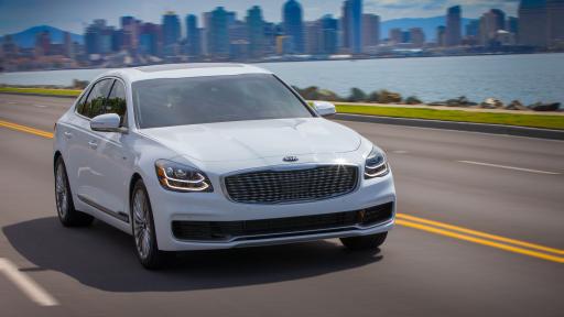 Exterior front end of KIA K900 from outside with a city in the background and sun shining as it rides on a highway.