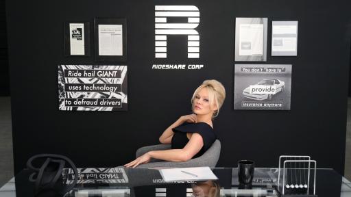 Pamela Anderson sitting in a chair at a desk with a black wall behind her.