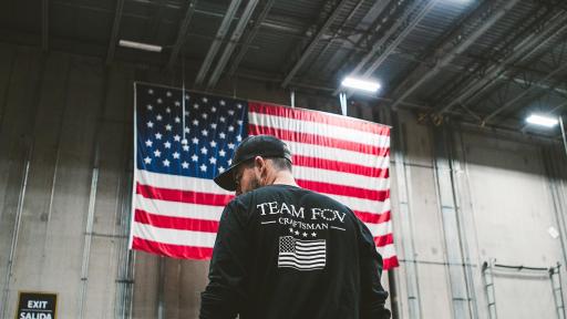 Brian Steorts standing in a factory with back to the screen, sporting a Team FOV Craftsman shirt. The American flag is on a wall in the background.