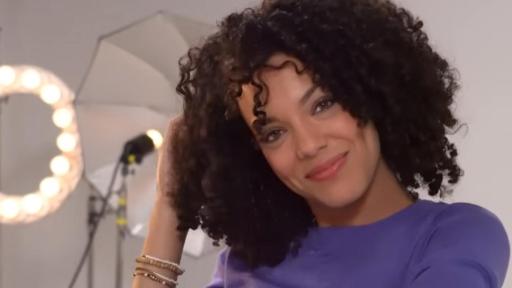 Woman with dark beautiful curly hair, wearing a purple shirt is looking at us.