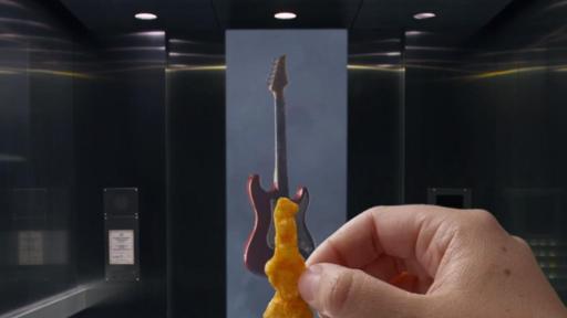 A hand holds a guitar-shaped Cheetos in front of a real guitar. Play the video.
