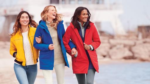 Three woman walking along the beach. Left wearing a yellow coat, middle in and blue coat, and right in a red coat.