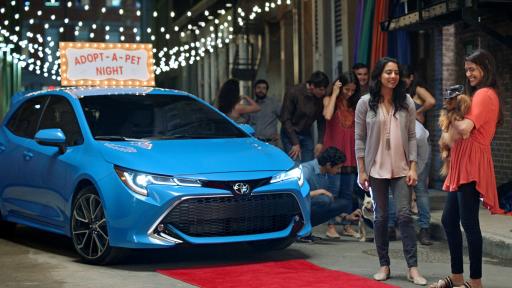 The front of a blue 2019 Toyota Corolla Hatchback on a small city street surrounded by people.
