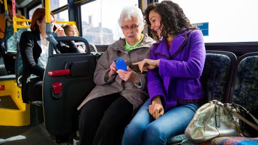 An elderly woman and younger woman sitting next to each other on a bus, pointing at the elderly woman's phone.