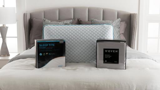 Bed with two packages on it, one is the sleep tite mattress protector and the other is woven sheet set.