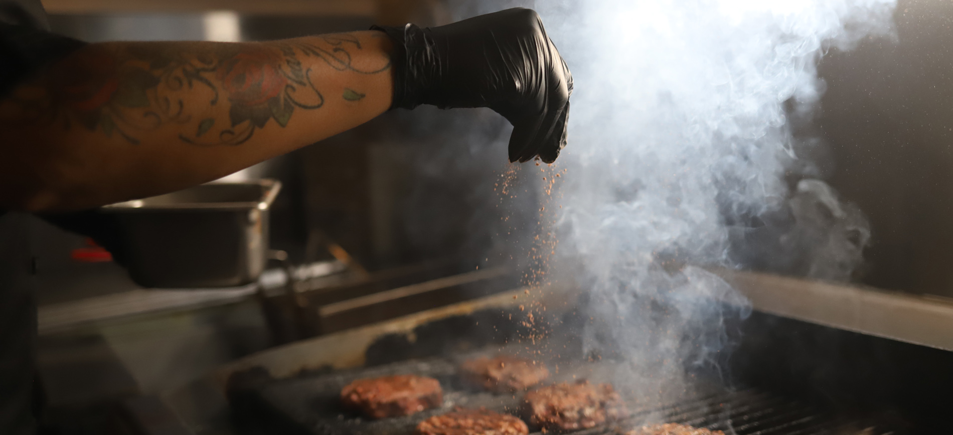 Person wearing gloves seasoning burger patties on a grill