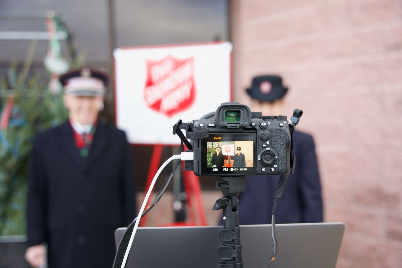 Camera shot of Commissioners Kenneth and Jolene Hodder standing next to a Salvation Army Red Kettle