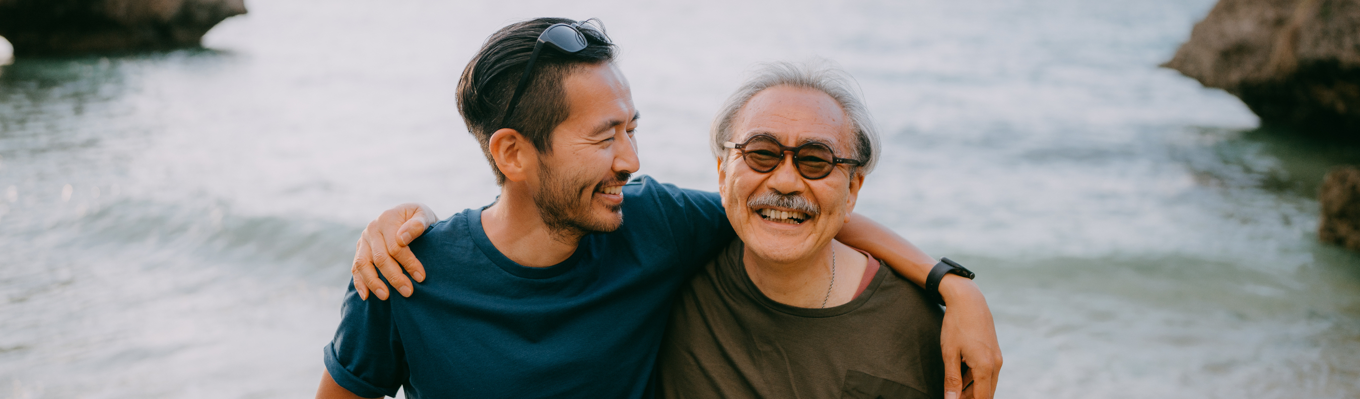 An elderly Asian father wearing glasses and a brown shirt and his adult son wearing sunglasses and a teal shirt smile side-by-side with their arms around each other. They are standing on a beach with the water and rocks in the background on a sunny day.