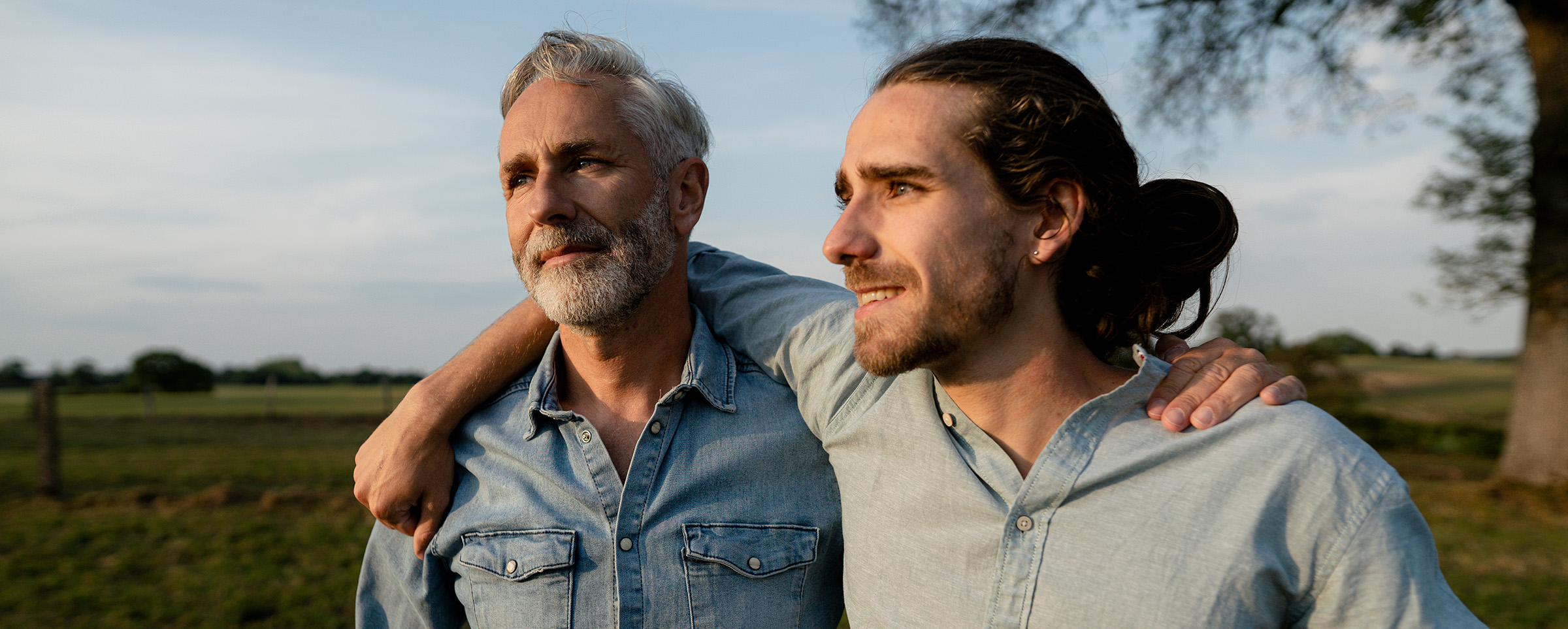 Two men stand side-by-side with their arms on each other’s shoulders outside with a field and tree behind them staring into the distance. The young man with long brown hair pulled back and a beard is wearing a blue button-down shirt. The older man with gray hair and a beard wears a jean button-down shirt.