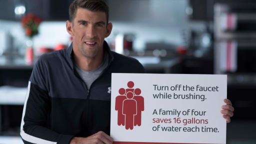 Michael Phelps holding up a sign that says "Turn off the water while brushing. A family of four saves 16 gallons of water each time."